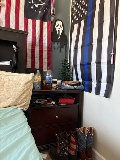 The image displays a bedroom interior. Various flags are hung on the wall, including a "thin blue line" American flag, and an American flag with the Assassin's Creed logo. A "Scream" mask decoration and a small Christmas tree are also on the wall. Bottled water, cowboy boots, and a dark wooden cabinet are present in the room. A beige pillow and light blue bedsheets are in the foreground.