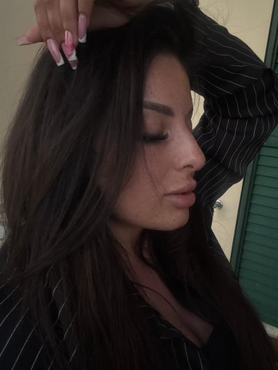 A woman with long, dark brown hair is lying down, resting her head on her hand. She is wearing a black and white striped shirt. The woman's eyes are closed, and she has freckles on her face. Her fingernails are manicured with a pink and white design. The background appears to be a green-colored surface, possibly a door or wall. The location in the city of Municipal Unit of Lefkada, Greece is not discernible.