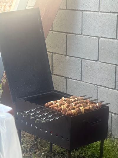 In Balivka, Ukraine, a top-down view captures a barbeque grill laden with skewers of meat and onions, ready for cooking. The grill, made of dark metal, rests on a paved area with a pattern of grey rectangular stones. To the right of the grill, green grass and dried branches suggest an outdoor setting, possibly a backyard or park. The sunlight casts a warm glow on the scene, indicating it is likely daytime. The meat on the skewers appears to be beef or lamb, interspersed with thick slices of onion. The grill itself has a segmented design with a series of circular openings, possibly for airflow or holding charcoal. The overall impression is one of preparation for a meal, evoking a sense of casual outdoor dining.