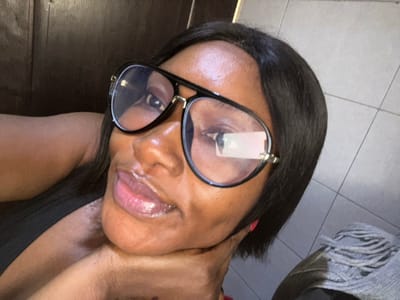The image displays a close-up portrait of a woman, taken indoors in Dzhidu, Nigeria. She has dark skin, dark, straight hair, and is wearing large, black-framed glasses with clear, reflective lenses that feature gold-colored accents. Her lips are glossy, and she looks directly towards the camera with a slight smile. Her right hand, showing a thumb with red nail polish, is positioned under her chin/jawline. She is wearing a dark-colored garment visible on her left shoulder and chest. The background is divided into two sections: a dark brown, possibly wooden, surface in the upper left, and a light grey tiled wall in the lower right. A grey, textured fabric object is partially visible in the bottom right corner of the frame.
