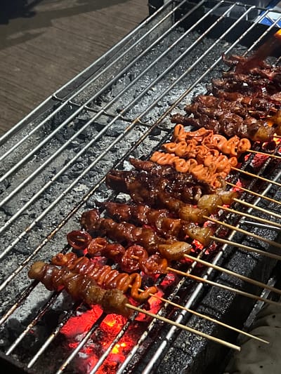 The image shows a close-up view of various meat skewers being grilled over hot coals. The skewers, made of small pieces of meat and some coiled intestine-like parts, are arranged on a metal grill. The coals beneath are glowing red, indicating intense heat and suggesting the grilling is in progress. The meat appears to be glazed, with some pieces having a dark, caramelized sheen.
The scene is set outdoors, likely at a street food stall given the rustic grill and the lighting which suggests it's evening or nighttime. The overall impression is one of a lively, casual food experience, common in places like Cuyo, Philippines, where grilled street food is popular. There is no visible text in the image. The focus is entirely on the appetizing presentation of the grilled meat, with the intense heat from the coals adding a dynamic element to the scene.