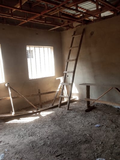 An interior view reveals an unfinished room. The walls are roughly plastered, with visible wooden scaffolding and a ladder propped against one wall. Two windows allow sunlight to stream in. The ceiling is a network of exposed wooden beams and metal sheeting. The floor is covered in dirt and debris, suggesting ongoing construction. Overall, the space conveys a sense of rawness and incompletion.