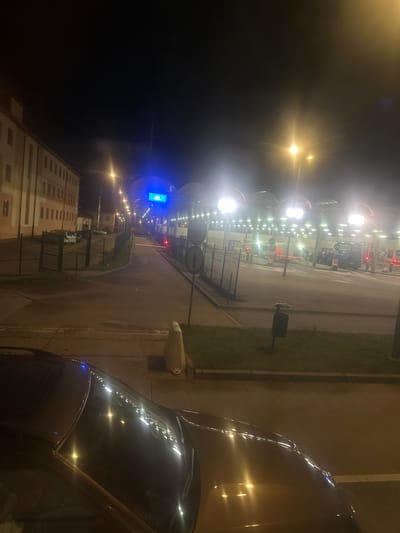 The image captures a night scene at the Hrebenne border crossing in Poland. A dark-colored car's hood and windshield are prominently featured in the foreground, reflecting the bright lights of the border post. In the background, a long, multi-lane structure with illuminated booths and overhead lighting signifies the border control area. Vehicles, including vans and cars, are visible in the queues. A blue digital sign with white text is mounted above the booths. To the left, a multi-story building with several windows is illuminated, suggesting adjacent facilities. The ground appears to be paved, with grassy areas and a trash receptacle visible on the right side of the road. A yellow concrete barrier is positioned on the right side of the image, near the foreground. The scene is lit by streetlights and the intense lighting of the border crossing.