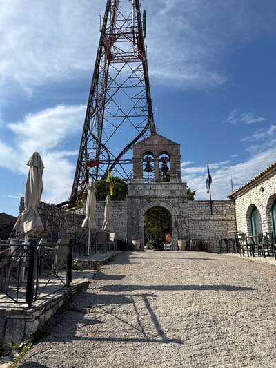 The image depicts an outdoor scene in what appears to be a public space with a stone building that resembles a church or historical structure. A tall, lattice-like metal tower, possibly a telecommunications tower or part of a structure, dominates the left side of the frame. In front of the stone building, there is a bell tower with two large bells. A flag, blue and yellow, is mounted on the building, and another small flag, possibly red, is visible on the metal tower. The ground is paved with gravel, and there are several tables and chairs, some with closed umbrellas, suggesting an outdoor seating area for a cafe or restaurant.

The time of day is suggested by the long shadows cast across the paved area, indicating late afternoon or early morning. The sky is a vibrant blue with scattered white clouds, suggesting pleasant weather. The architecture of the stone building, with its arched windows and stone blocks, points to a location with a historical or Mediterranean feel, consistent with the provided location context of Greece. No people are visible in the frame. There is no legible text in the image. The overall impression is one of a quiet, possibly tourist-oriented location on a clear day.