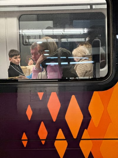 The image provides a detailed view into the interior of a West Midlands Railway passenger train, viewed from outside through a window, indicating its operation within the Metropolitan Borough of Solihull, United Kingdom. The train's exterior beneath the window features a dark purple base with scattered orange diamond patterns.
Inside the train, four individuals are visible. On the far left, a young male, possibly a teenager, with short dark hair, wears a black tracksuit top with white arm stripes. He looks forward and slightly left, with his mouth slightly ajar, his right hand resting on a flat, light-colored object. Adjacent to him, an adult male with short grey hair and a light blue top looks towards the right, holding a small, multi-colored object in his right hand. Next to him, an adult female with blonde hair and glasses wears a dark jacket over a light pink garment, her right hand supporting her chin as she looks downwards and to the right. A silver bracelet is visible on her left wrist. Partially obscured to the right of her, another adult female with blonde hair is seen from the back, wearing a quilted beige jacket and looking downwards. Overhead, multiple fluorescent strip lights illuminate the train's cabin. Reflections are visible on the window surface, slightly obscuring the interior.