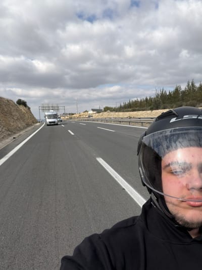 The image depicts a highway scene in Monforte del Cid, Spain, taken from the perspective of an individual wearing a motorcycle helmet. The individual's lower face, including the nose, mouth, and chin, is visible on the lower right, partially obscured by a clear visor of a black LS2 full-face helmet. The person has fair skin and a slight pouting expression, wearing a black hoodie or jacket.
The view extends forward along a multi-lane asphalt highway with white solid lines marking the left shoulder and white dashed lines indicating travel lanes. A metal guardrail runs along the right side of the road. In the distance, a white recreational vehicle (RV) or campervan is visible in the left lane, followed by a smaller, dark-colored vehicle, both travelling away from the viewpoint. An overhead gantry structure spans the highway further down.
To the left of the highway, an excavated hillside with exposed reddish-brown soil and sparse vegetation is present. To the right, beyond the guardrail, lies a stretch of disturbed earth, followed by a dense treeline of evergreen trees. In the far background on the right, faint structures and a tall communication mast are discernible. The sky is partially cloudy, featuring a mix of white and grey clouds with interspersed patches of blue.