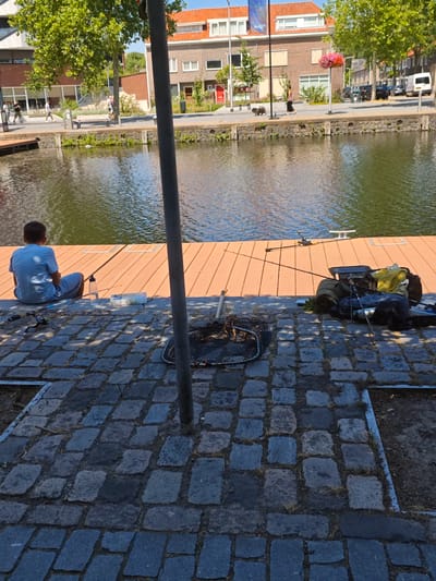 A person is fishing by a canal in Tilburg, Netherlands. The scene depicts a person sitting on a wooden dock facing the water, with fishing equipment nearby. The canal is calm and reflects the surrounding buildings and trees. The area is paved with cobblestones in the foreground, and the background shows residential buildings and trees. The weather appears to be sunny and warm.