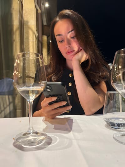 A young woman is sitting at a table in an outdoor setting, possibly in Polignano a Mare, Italy, based on the appearance of the stone architecture and dark sky. She is resting her head on her left hand, holding a black smartphone in her right hand. The woman has brown hair, fair skin, and is wearing a black top with gold buttons.
The table is covered with a white tablecloth. Several wine glasses are on the table; one is in the foreground. The background shows a stone wall and a dark sky. The scene is lit by artificial light. The woman is the primary subject, her focus seems to be on the smartphone.