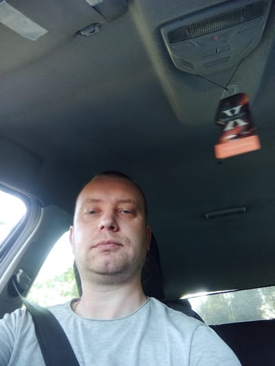 A bald white man wearing a grey t-shirt and a black seatbelt is sitting in the driver's seat of a car. He is looking directly at the camera. The interior of the car, including the dashboard, roof, and a car air freshener, is visible. The image was taken from inside the car, looking forward. The background outside the car appears to be a natural environment with trees. The location is Буйніцкі сельскі Савет, Belarus.