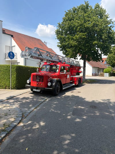 A red Deutz ladder truck, identified by the license plate "IN-S 112", is parked on a residential street in Ingolstadt, Germany. The truck features a large, extendable ladder mounted on its roof and has "Feuerwehr 112" written on its side. The truck is positioned on the right side of the road, which is paved with asphalt. To the left of the truck, a neatly trimmed green hedge runs along the sidewalk. A blue traffic sign with symbols of a pedestrian and a bicycle is visible next to the hedge. In the background, a white house with a red tile roof is situated behind the hedge, and a large green tree with dense foliage stands behind the truck. The sky is blue with scattered white clouds. The scene is illuminated by sunlight, casting shadows from the trees onto the street and sidewalk.