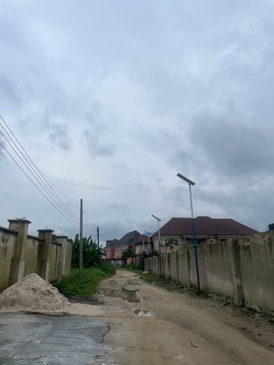 The image depicts an overcast sky above a dirt road in Elimgbu, Nigeria. The road is uneven and has several large puddles from recent rainfall. On the left side of the road, a tall, beige concrete wall topped with spikes runs along the property line. Next to the wall is a large pile of light-colored sand. Behind the sand pile, there is a dense growth of green vegetation. Power lines are strung across the upper left portion of the image, supported by a utility pole.

Further down the road on the left, a line of houses with dark roofs is visible. On the right side of the road, a similar beige concrete wall extends into the distance, also with a rough texture. Several modern streetlights with solar panels are spaced along this wall. The houses visible on the right are multi-story buildings with reddish-brown roofs and light-colored facades. The overall scene suggests an urban or suburban environment under cloudy conditions.