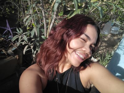 A young woman with reddish-brown hair is taking a selfie outdoors. She is smiling with her eyes closed, and her mouth is open, revealing her teeth. She is wearing a black tank top and a necklace. The background consists of various plants and foliage, with some parts appearing out of focus. Sunlight casts shadows on the ground and on her skin. The overall scene suggests a casual, outdoor setting, possibly in a garden or natural area. The location is identified as Punta de Piedras, Venezuela.