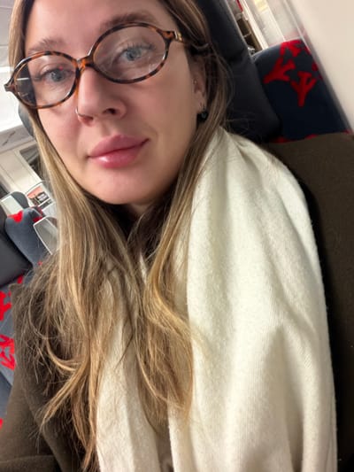 A woman is depicted in a close-up selfie taken within a public transport vehicle in Pavlovsk, Russia. She has light-colored eyes, full lips, and a nose piercing (nostril ring). She wears tortoise-shell oval-shaped eyeglasses and a dark stud earring in her right ear. Her long, light brown hair frames her face and extends over her shoulders. She is dressed in a dark, possibly brown, coat or jacket, and an off-white scarf is draped over her left side. Behind her are visible passenger seats characteristic of a train or bus, featuring dark grey upholstery with a repeating red pattern. A seatbelt buckle is partially visible on the seat adjacent to her. The scene suggests an indoor environment.