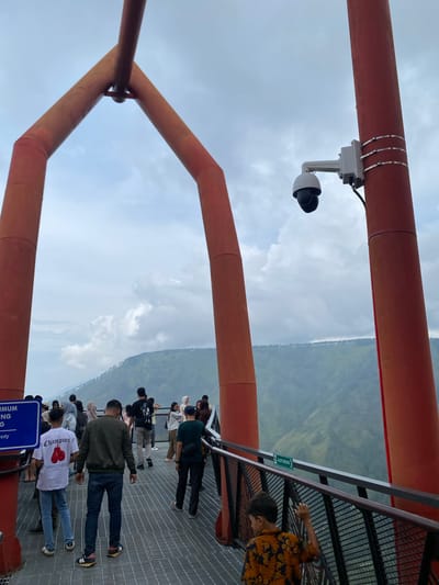 The image captures a group of people standing on a metal grate walkway, likely part of an elevated structure or observation deck, overlooking a mountainous landscape. Dominating the foreground are two large, curved, bright orange support beams, suggesting a unique architectural feature, possibly an entrance or part of a bridge. A security camera is mounted on one of the orange beams, facing outward.

In the midground, several individuals are dispersed across the walkway. Some are looking out at the vista, while others are engaged in conversation or walking. The people are dressed in casual attire, including t-shirts, jackets, and pants. One person in the foreground is wearing a white t-shirt with a graphic of boxing gloves, and another is wearing a dark green jacket and jeans. A young boy in an orange and black patterned shirt is visible towards the right, leaning on the railing.

The background reveals a vast, verdant valley with rolling hills and mountains shrouded in a light mist, indicative of a natural scenic location. The sky is overcast with scattered clouds. A green sign with white text, possibly an evacuation route indicator, is affixed to the railing of the walkway. The overall impression is a public viewpoint offering panoramic views of natural scenery. The location is identified as Turpuk Malau, Indonesia.