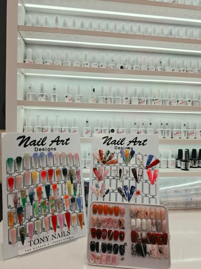 This image captures a brightly lit display of nail products and art designs in what appears to be a nail salon or beauty studio, identified as "TONY NAILS THE BARBER & LASHES STUDIO" located in Mladá Boleslav, Czechia.

The central focus is a vertical display unit filled with rows of small white bottles, prominently labeled "gellak," suggesting gel nail polish. The bottles are neatly organized in illuminated shelves, with visible numbers like 205, 207, 208, 265, 268, 273, 274, and others.

In the foreground, there are three main displays of nail art. Two rectangular boards are titled "Nail Art Designs" in a stylized script. These boards showcase various sample nail tips with intricate patterns, colors, and textures. The lower board, bearing the "TONY NAILS" branding, features a gradient of colors, glitter, and abstract designs, each numbered and often accompanied by a numerical code (e.g., "50+002", "38+004", "47+006+010"). The upper display board, partially obscured, presents more elaborate and artistic designs, including animal prints, geometric patterns, and embossed details, also numbered (e.g., 17, 18, 20, 23, 24, 26, 27, 28, 29, 39, 40).

To the right, a white plastic tray holds numerous individual false nails, demonstrating a wide range of finished designs. These include solid colors, glitter, ombré effects, marbled patterns, and artistic embellishments, with one notably featuring the word "LOVE." The setting is well-lit with artificial light, creating a clean and inviting atmosphere for customers to browse the available options for nail services. No people are visible in the frame.