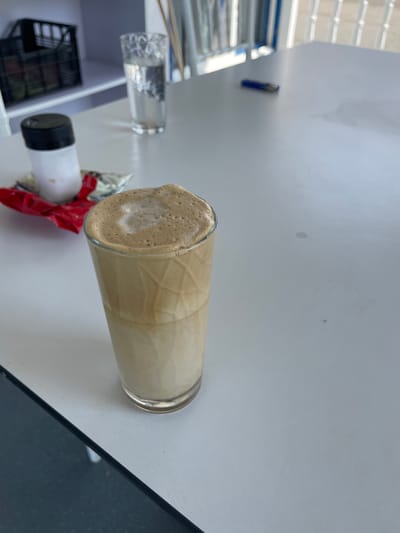 In Skala Avdiron, Greece, a table is set with a tall glass of frothy coffee, a glass of water, a black-capped white bottle, and a red wrapper. The glass of coffee, a Dalgona, displays distinct layers of light brown foam atop a pale beige liquid. Beside it, a clear glass holds water, and a salt shaker with a black lid stands next to a red foil wrapper. The tabletop appears to be a white laminate surface, reflecting light from overhead fixtures. In the background, a black plastic crate is visible on a shelf, and a blue pen rests on the table further back. The environment suggests a cafe or an outdoor seating area, with glimpses of white railings in the distance.