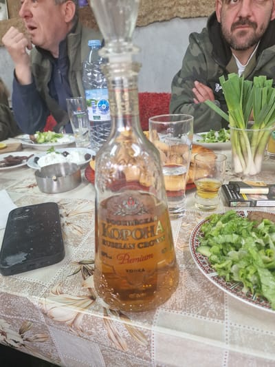 The scene presents a table setting with food, beverages, and personal items, likely outdoors. A bottle of "Korona" Russian Crown Premium Vodka is centrally located, near empty glasses, a plate of salad, a container of leeks, and a pack of cigarettes. Two men are partially visible; one is on the left side near a bottle of water, and another man, wearing a camouflage jacket, is visible near the leeks. A black smartphone sits to the lower right of the vodka bottle. The tablecloth has a floral pattern. There is an ashtray near the bottle of water, and plates with appetizers/salads. The background suggests an outdoor setting. The location, Vinarovo, Bulgaria is speculative.