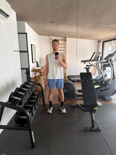 A man with short brown hair and a beard, wearing a white sleeveless shirt, blue shorts, black socks, and white sneakers, stands in a gym and takes a selfie in a mirror. He is holding a black smartphone in his right hand. The gym has a wooden wall ladder, a rack of dumbbells, a treadmill, an elliptical machine, and a weight bench. The flooring is black rubber mats. In the background, through a large window, palm trees and a glimpse of the sky are visible. The environment suggests a location in the city of Montuïri, Spain.
