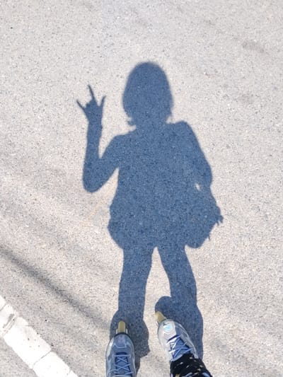 The image shows a shadow of a person on a paved surface, likely a road or sidewalk, in Ban Patan, Thailand. The person's shadow is casting a rock-on hand gesture, and they appear to be wearing rollerblades. The shadow's posture suggests they are standing still. The environment is outdoors, with the shadow indicating a sunny day. The paved surface is gray and textured, with some white markings visible on the left side, possibly a pedestrian crossing.