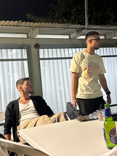 The image depicts a nighttime scene at a covered patio or outdoor seating area in Kfar Yona, Israel. Two young adult males are present.
The male on the left is seated, wearing a white tank top, a black cardigan, and light-colored cargo pants. He has short dark hair with a fade and is looking upwards and to his right (viewer's left). A silver chain with a pendant is visible around his neck. He is positioned behind a white table.
The male on the right is standing, partially obscured by the white table. He wears a light yellow t-shirt with a graphic print, dark pants, and eyeglasses. His hair is short and dark with a fade. He has a wristwatch and a silver bracelet on his left wrist and is holding a black, hard-shell case with a silver handle in his right hand. He is looking forward and to his right (viewer's left).
On the white table in the foreground, there is a green glass bottle of Maccabi beer, two clear plastic cups (one containing snacks like nuts, the other empty), a crumpled piece of silver foil, and a dark, rectangular object.
The background features large windows or glass panels with white, vertically-striped curtains. Above, a white corrugated roof structure or pergola is visible, from which a spherical, woven light fixture hangs. Dark foliage from trees is visible against the night sky above the roofline. The area is illuminated by artificial light sources.