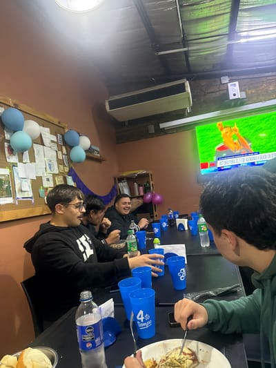 The image captures a casual indoor gathering of several young men in Sarandí, Argentina, focused on a large flat-screen TV. The scene is set around a dark table, laden with food and drinks. In the foreground, a man in a green hoodie eats from a white plate using a fork and knife, while another man in a black hoodie with white text is visible, both looking towards the TV. Further down the table, other men are seen, some laughing, contributing to a relaxed and social atmosphere.

The TV screen prominently displays Spanish text, "HISTORIA PURA: EL 7-1 DE ALEMANIA A BRASIL EN SEMI" (Pure History: The 7-1 of Germany against Brazil in the Semifinals) and "MASK 2014," indicating they are watching a recap or documentary about the infamous 2014 FIFA World Cup match. Blue plastic cups, some branded with "4to tiempo" (4th time/quarter, a sports term), are arranged on the table alongside water bottles, one clearly showing "PEPSI" and "500" (likely ml), and another with "70." A white plate holds a meal, possibly milanesa with fries, and bread rolls are in a metal bowl.

The room features a warm-toned wall, a bright circular light fixture, and an industrial-style ceiling with green tarp-like material. Blue, white, and light blue balloons decorate a wooden cork board covered with papers, while two purple balloons are on a shelf in the background, suggesting a celebratory or special occasion. An air conditioning unit is also visible. The overall mood is one of shared entertainment and camaraderie.