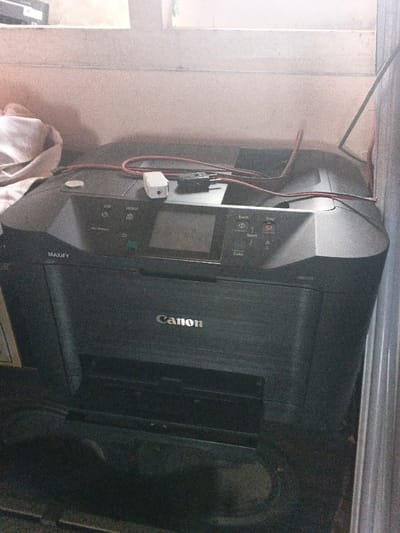 The image depicts an indoor scene in the city of Dogsenang Gorphoog, Bhutan. A black Canon MAXIFY MB5370 multifunction printer, identified by "Canon" and "MAXIFY" branding, is centrally positioned. Its control panel, featuring various buttons (e.g., Power, Home, Back, Start, Stop, Color) and an unlit LCD screen, is visible on the top surface. Resting on the printer's top are a white USB wall charger/adapter, a black two-pronged electrical plug, and a loosely coiled red cable. To the left of the printer, a crumpled light-colored fabric object is partially visible. The background consists of a light-colored wall, possibly with an unseen window above providing diffused light. A vertical metallic or plastic trim piece and a dark cable run along the right edge of the frame. No individuals are present.