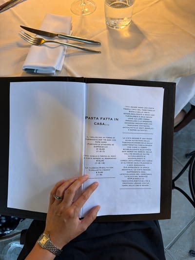 This image captures a close-up of a table setting at a restaurant, with a focus on a menu and a person's hands. The setting appears to be outdoors, with natural light suggesting daytime. The menu, presented on a white sheet of paper bound in a dark cover, features Italian text describing pasta dishes and a breed of pig. Visible text includes "PASTA FATTA IN CASA...", "I PICI SENESI SONO UNA PASTA", "LA CINTA SENESE È UNA RAZZA SUINA AUTOCTONA...", and prices in Euros.

A person's hands are visible, one holding the menu and the other resting on the table. The person is wearing a wristwatch and a ring. The table is draped with a light-colored cloth. A white napkin is folded neatly, accompanied by a fork and a knife. A partially visible glass with bubbles suggests a beverage. In the background, a dark chair and a glimpse of what appears to be a tiled floor hint at an outdoor or semi-outdoor dining environment. The overall impression is one of an intimate dining experience, focusing on traditional Italian cuisine. The location context is Asciano, Italy.