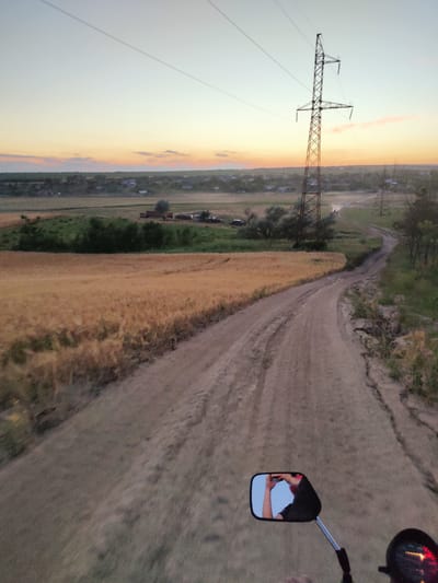 This is a point-of-view photograph, likely taken from a motorcycle, looking down a dusty rural road. The sun is setting, casting a warm, golden light across a field of ripe wheat to the left. In the distance, a small village with scattered houses is visible, nestled amongst trees and fields. Power lines stretch across the sky, supported by tall lattice towers, indicating an electrified rural area. The sky itself displays the soft hues of twilight, with hints of orange and pink near the horizon and a pale blue overhead.

The road is unpaved, with tire tracks suggesting recent use by vehicles. The overall setting is peaceful and agrarian, characteristic of the Moldovan countryside. In the foreground, a motorcycle's rearview mirror is visible, reflecting the rider holding a phone and taking the photograph. The rider's arm and part of their black shirt are also visible. To the right of the mirror, a portion of the motorcycle's instrument cluster can be seen, with a red indicator light illuminated. The time of day is clearly evening, with the low sun and pastel sky creating a serene atmosphere. There is no visible text in the image.