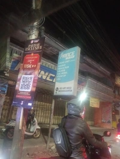 This media file depicts a nighttime scene on a street in Calukan RT 1 RW 5 Keboansikep, Indonesia. The primary subjects are two people on motorcycles, though one is more prominently featured. The person in the foreground is wearing a dark leather jacket and a helmet, with their back to the camera, as they sit on a motorcycle. Another motorcycle, light-colored, is parked to the left, with its rider obscured.
The setting is a street with buildings in the background, suggesting a commercial area. Several banners and signs are visible, indicating various promotions and services. One banner, in maroon and black, advertises a "PAYDAY EXCLUSIVE" event from KAKKOII SIDOARJO, offering special prices of 115K++ and 115K+F, and directs users to scan a barcode for reservations during the period of June 29th to July 3rd, 2026. Another sign, in light blue, promotes "PRINCE MOBIL" and lists services such as "HEAD UNIT (ANDROID), AUDIO, KACA FILM, ALARM, AC MOBIL, TUNE UP, DLL," along with contact numbers. A yellow sign with text that is partially legible mentions "GANTI OLI" (oil change) and "INGERSTTEEL."
The time of day is clearly night, as indicated by the darkness and the artificial lighting from streetlights and building interiors, which create strong highlights and shadows. The overall atmosphere is that of a busy street at night, possibly near shops or service centers. The presence of a "HALAL" sticker on one of the banners suggests a cultural context. The street appears to be paved, and there are electrical wires overhead, typical of urban or semi-urban environments.