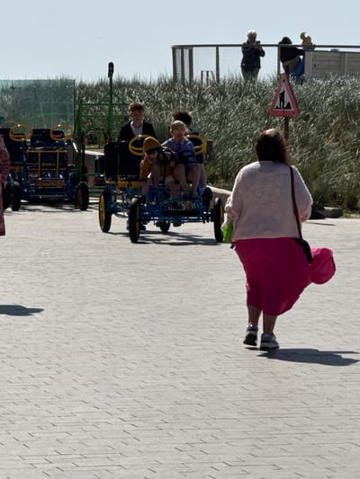 This vertically oriented image captures a bright, sunny outdoor scene in Middelkerke, Belgium, depicting a paved promenade bordered by dense, dune-like vegetation.
In the immediate foreground, a person with dark hair, viewed from behind, walks along the light gray, textured paved path. They are wearing a light-colored, possibly fluffy, top or jacket and a striking bright pink skirt or dress. Their elongated shadow, along with others visible on the path, indicates the time of day is either mid-morning or late afternoon under clear skies.
Further down the path, several individuals are engaged in recreational activities, primarily riding pedal karts. Two children or young adults are seated in a blue pedal kart with yellow steering wheels and accents. Another person wearing a black jacket is visible on a separate pedal kart. Additional figures are present in the background, including one person with white hair, some appearing to also be on karts or walking.
To the left, a metal fence runs parallel to the promenade, separating it from a bushy expanse of tall, light green grass or reeds, consistent with coastal dune flora. A pink-bordered, triangular warning sign is affixed to the fence, displaying a silhouette of a person pushing a cart, likely indicating a path for similar recreational vehicles or specific pedestrian usage. The overall atmosphere suggests a leisurely day in a coastal recreational area. No discernible text is visible on the sign or elsewhere in the image.