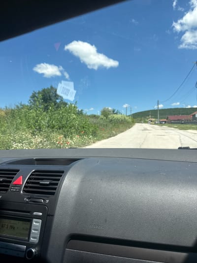 The image shows a view from inside a vehicle, looking through the windshield. The dashboard of the car is in the foreground, with the stereo system and air vents visible. Outside the car, a paved road leads into the distance, lined with green vegetation and wildflowers. In the background, there are houses and hills under a clear blue sky with scattered clouds. Two small stickers with QR codes are attached to the windshield. The scene appears to be a rural or semi-rural area. Based on the provided context, this location is in the city of Suvorovo, Bulgaria.