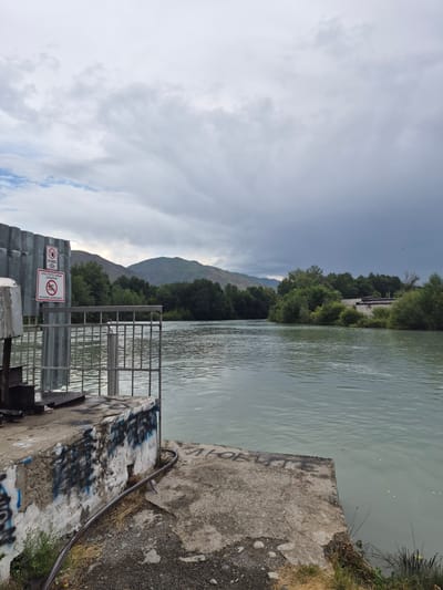 The image depicts a view of a river with a concrete embankment in the foreground. On the embankment, there is some graffiti and a metal railing. Behind the railing, the river flows, with trees and hills in the background under a cloudy sky. Two signs with prohibition symbols are attached to a metal fence on the left side of the image. The sign on top shows a hand with a line through it, likely indicating "no touching". The lower sign shows a figure swimming with a line through it, indicating "no swimming". The text on the signs appears to be in Cyrillic script, suggesting the location is in a region where this script is used, consistent with Tekeli, Kazakhstan. The overall scene suggests an area near a river where access might be restricted or dangerous for certain activities.