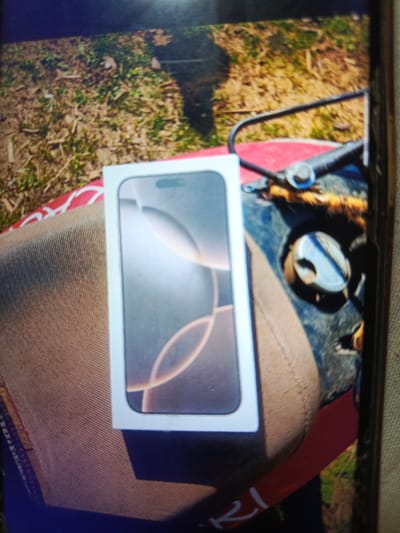 An iPhone 14 Pro box is placed on a red object, which appears to be part of a vehicle, possibly a motorcycle. The vehicle has a dark, metallic component with a circular gauge and some frayed orange material visible. The scene is outdoors, with dry grass and leaves on the ground. The background is blurred but suggests a natural, possibly rural environment. There are no people visible in the image.