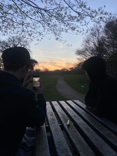 Two people are sitting on a weathered wooden bench in a park at dusk or dawn. One person, wearing a dark hooded sweatshirt and a baseball cap, is holding a smartphone and taking a photo of the sunset or sunrise visible through the trees. In the background, a path leads into the distance through a grassy area. The setting is likely in Broxbourne, United Kingdom, given the prompt. A small, green object sits on the bench. The other person, wearing a dark hooded sweatshirt, is sitting next to the photographer. Bare branches of trees are visible against the sky.