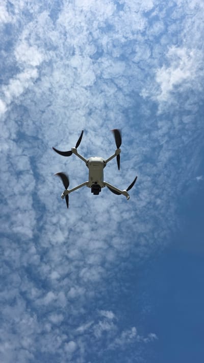 A drone is flying in a blue sky with white, puffy clouds. The drone is white with black rotors and a camera is visible. The location cannot be determined as Valchi Dol, Bulgaria based on the image.