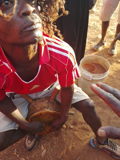 A man with curly hair and a red shirt plays a wooden drum while seated. His hands firmly grip the drum's surface. Beside him, a white bucket filled with a brownish liquid sits on the ground. The ground is reddish-brown and looks like dirt. In the background, some people are standing. The image is brightly lit with some areas of the scene appearing overexposed.