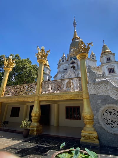 A low-angle shot showcases a Buddhist temple in Dĩ An, Vietnam. The temple exhibits a white and gold color palette. Golden pillars with decorative elements flank the temple's entrance. The temple is surrounded by a clear blue sky, a tree, and a tile pathway.