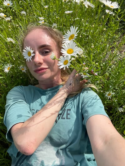 A young woman with blonde hair and daisies over her eyes lies in a field of daisies and tall green grass. She is wearing a teal t-shirt with a faded graphic on the front. Her right arm is bent, resting on her chest, and her left arm is outstretched to the side. The scene is lit by bright sunlight, casting shadows from the grass and flowers onto the woman and her clothing. The background is a blur of green grass and white daisies, suggesting an outdoor, natural setting, likely in the city of Yasnoe, Russia, given the context.
