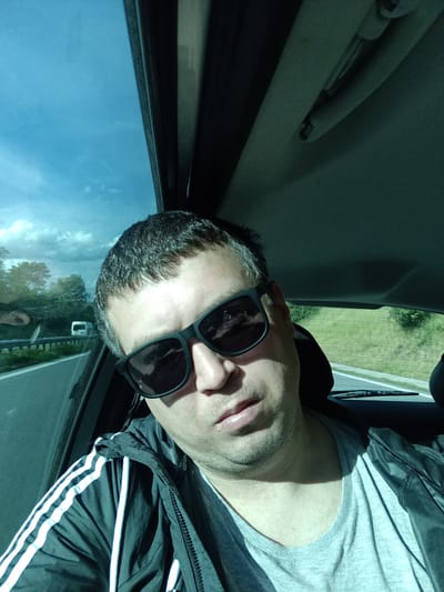 A daylight selfie captures an adult male seated as a passenger in a moving vehicle, located within or near Třemošnice, Czechia. The subject, appearing to be in his 30s or 40s, has short dark hair with visible grey streaks and light facial stubble. He wears black-framed sunglasses, a grey crew-neck t-shirt, and a black zip-up jacket with three white stripes on the sleeves. His head is tilted slightly, and he looks directly at the camera. Sunlight enters from the left, illuminating the left side of his face and upper body while casting shadows on the right. Through the car's left window, a multi-lane highway, a white van in the distance, a metal guardrail, green foliage, and a blue sky with white clouds are visible. The vehicle's dark interior headliner, a sun visor mechanism, and the rear window with defroster lines are also discernable.