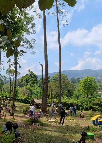 This image depicts a lively outdoor playground set amidst a lush, green landscape. Several tall, slender trees dominate the foreground, their branches reaching towards a bright blue sky dotted with fluffy white clouds. In the background, rolling hills covered in dense foliage create a picturesque backdrop. The time of day appears to be late morning or early afternoon, given the clear and sunny weather.
The playground itself features a variety of attractions. A swing set is visible on the left, with a few children seated on it. In the center, a circular spinning ride with colorful seating is occupied by several people, including children and adults. Another similar spinning ride is located further to the right. Nearby, a small, rustic wooden hut offers shelter. Brightly colored playground equipment, including yellow and blue slides, can be seen on the right side of the frame.
Various people are scattered throughout the playground, enjoying the recreational facilities. Some are actively engaged in playing, while others appear to be watching or resting. The overall atmosphere seems cheerful and relaxed, suggesting a pleasant outing for families and visitors. The setting, identified as Laugumba, Indonesia, hints at a tropical or subtropical environment, evident from the abundant greenery and tall trees. No visible text is present in the image.