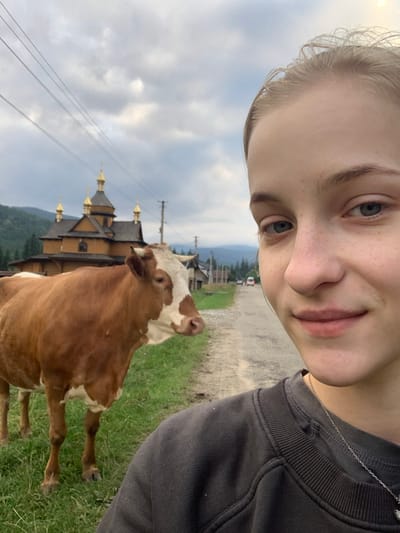 A selfie-style image depicts a rural mountainous scene in Vorokhta, Ukraine. The foreground on the right is occupied by the partial view of a young female with fair skin, light hair, and blue eyes, presenting a slight smile while wearing a dark gray sweatshirt and a silver chain necklace. To her left, a brown and white cow with horns stands on green grass, facing towards the right.

In the midground, the Church of the Dormition of the Theotokos is visible; it is a traditional wooden Eastern Orthodox church with five prominent golden domes, each topped with a cross, situated on a slight elevation. A dirt or gravel road extends from the mid-right into the background. Along this road, rural buildings are visible, and in the far distance, a white vehicle (possibly a van) and another dark vehicle are present. Utility poles with multiple power lines stretch across the upper left and center of the frame. The background features dense green forested mountains under an overcast sky with significant cloud cover.