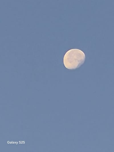 A crescent moon is visible in a clear, light blue sky. The moon is illuminated from the right, with its darker portion facing left. A watermark reading "Galaxy S25" is present in the lower-left corner of the image. The scene is observed in Pątnów, Poland.