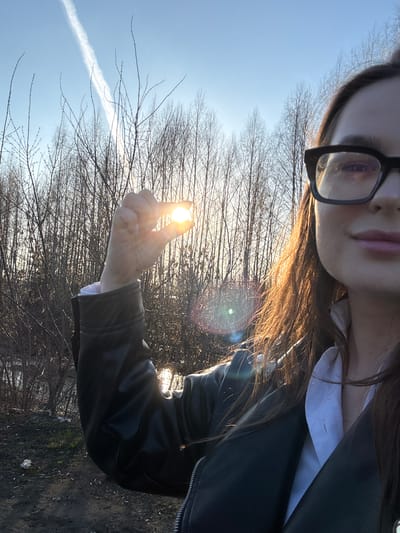 This media file captures a close-up, playful outdoor selfie, likely taken during either sunrise or sunset due to the low angle and golden glow of the sun. The subject is a person, appearing to be female, with dark hair cascading over her shoulder. Her face is partially visible in the upper right corner, revealing dark-rimmed glasses, a portion of her eye, and pink-toned lips. She is dressed in a white collared shirt and a black leather jacket.

Her right arm is extended, with her hand positioned so that she appears to be playfully "holding" the bright, radiant sun between her thumb and forefinger. This creates a strong focal point and a significant lens flare effect.

The background features a clear, bright blue sky with a distinct white airplane contrail stretching across the left side. Numerous bare tree branches, indicative of late autumn, winter, or early spring, frame the sky and the glowing sun. The ground beneath the trees is visible in the lower portion of the image, appearing as earthy terrain with sparse, dry vegetation and some small pieces of debris. The overall impression is one of a bright, clear day in an outdoor setting in Novoe Mart'yanovo, Russia. There is no visible text in the image.