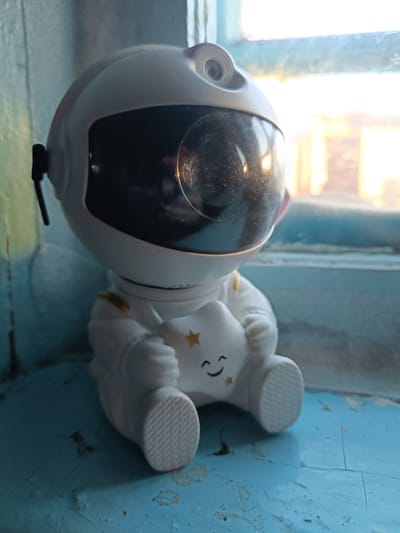 A close-up, eye-level shot shows a white astronaut figurine with a black visor, sitting on a cracked blue painted surface. The figurine is holding a white star with a smiley face and small yellow stars. The background consists of a window with sunlight filtering through, casting a warm glow on the scene. The setting appears to be indoors, possibly a windowsill. No specific location in Balakhta, Russia can be determined from the image.
