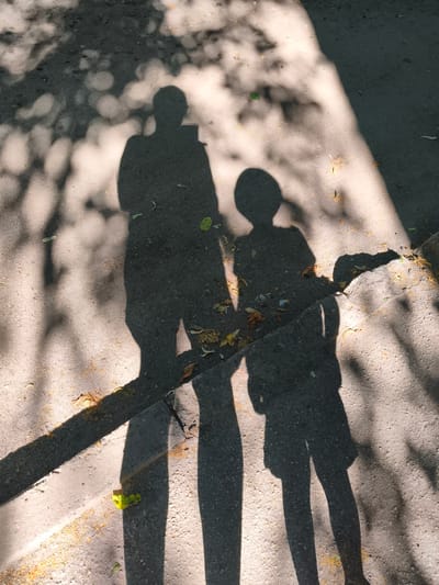 This media file shows the shadows of two people cast on a textured, paved surface, likely outdoors. The shadows suggest one person is taller and perhaps an adult, while the other is shorter, possibly a child. The taller shadow appears to be holding a rectangular object, which could be a phone. Both individuals' full bodies are depicted, from head to toe. The ground beneath the shadows is a mixture of asphalt and concrete, with visible debris such as dried leaves and small yellow flower petals scattered across it. Some green leaves are also present, along with a bright yellow object, possibly a toy or a flower, near the edge of the paved area.
The scene is illuminated by dappled sunlight filtering through trees, creating a pattern of light and shadow across the ground. The strong shadows indicate that the sun is high in the sky, suggesting it is midday. The clear shadows and the bright illumination imply fair weather, likely sunny. The presence of fallen leaves and petals hints at it being autumn or late spring. The setting appears to be a public space, possibly a park, a sidewalk, or a pathway in Sokolivske, Ukraine. The shadows themselves evoke a sense of companionship and shared experience. No visible text is present in the image.