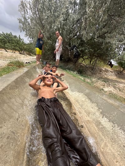 In Kan Mehtarzai, Pakistan, a group of boys slide down a concrete aqueduct filled with water. The aqueduct is located outdoors, surrounded by dry, rocky terrain and sparse vegetation, with trees visible in the background. Two men stand at the top of the aqueduct, observing the children. One man, wearing a dark t-shirt and yellow shorts, points upwards. The other man, in a white tank top and white shorts, stands next to him. The children are dressed in swimwear, with one boy in the foreground wearing dark, wet trousers. The sky is overcast, suggesting a warm, possibly humid day. The scene appears to be a recreational activity, possibly taking advantage of the water flow in the aqueduct on a hot day.