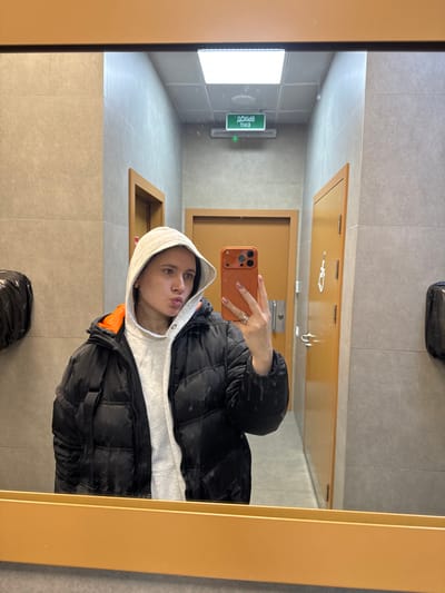 A person in a black puffer jacket and grey hoodie takes a selfie in a mirror. The setting appears to be a public restroom with grey tiled walls and a yellow-gold framed mirror. Reflected in the mirror are bathroom fixtures, a door, and a sign that includes the words “EXIT” in green lettering. A black bag is also visible in the reflection on the floor.