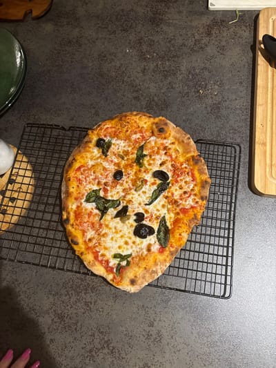 The image, captured in a kitchen in Ness Ziona, Israel, displays an oblong-shaped pizza centered on a black wire cooling rack. The pizza features a golden-brown crust, melted mozzarella cheese, visible tomato sauce, scattered green basil leaves, and several black olives. The cooling rack rests on a dark gray, textured countertop. In the upper left background, a portion of a dark green glazed plate and a light-colored wooden trivet or stand are partially visible. To the upper right, a section of a light wooden cutting board with a dark kitchen utensil (possibly tongs) is present. The lower-left corner of the frame includes the fingertips and nails, painted pink, of a person's left hand.