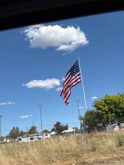 The image depicts an American flag flying on a flagpole against a bright blue sky with scattered white clouds. The flag is positioned to the right of the frame, unfurling in the wind. In the foreground, dry, tall grass is visible. Behind the grass, a chain-link fence runs horizontally across the image. Beyond the fence, several white RVs or trailers are parked in what appears to be a storage lot, with a few trees and streetlights also present. A dark, blurred object runs across the top of the image, possibly indicating the perspective is from inside a vehicle. The scene is outdoors during the daytime.