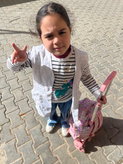 A young female child stands outdoors on a light-colored paved surface, specifically interlocking concrete pavers arranged in a herringbone pattern. The scene is illuminated by direct sunlight, casting distinct shadows. The child, appearing approximately 5-8 years old, has dark hair pulled back and looks directly at the camera with a neutral expression. She is dressed in blue denim jeans, light-colored sneakers, a striped long-sleeved shirt featuring a teal unicorn graphic, and an unbuttoned white long-sleeved smock or coat. Her left hand is raised, forming a V-sign. Her right hand grasps the extended pink handle of a multi-patterned pink rolling backpack positioned on the ground beside her right foot. The location is Ksar-el-Kebir, Morocco.